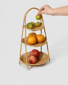 Hand placing fruit on the upper tray of Aina’s three-tier faux rattan fruit stand