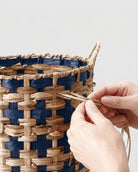 Hands weaving natural grass strands into the deep navy and natural two-tone pattern of Iris basket
