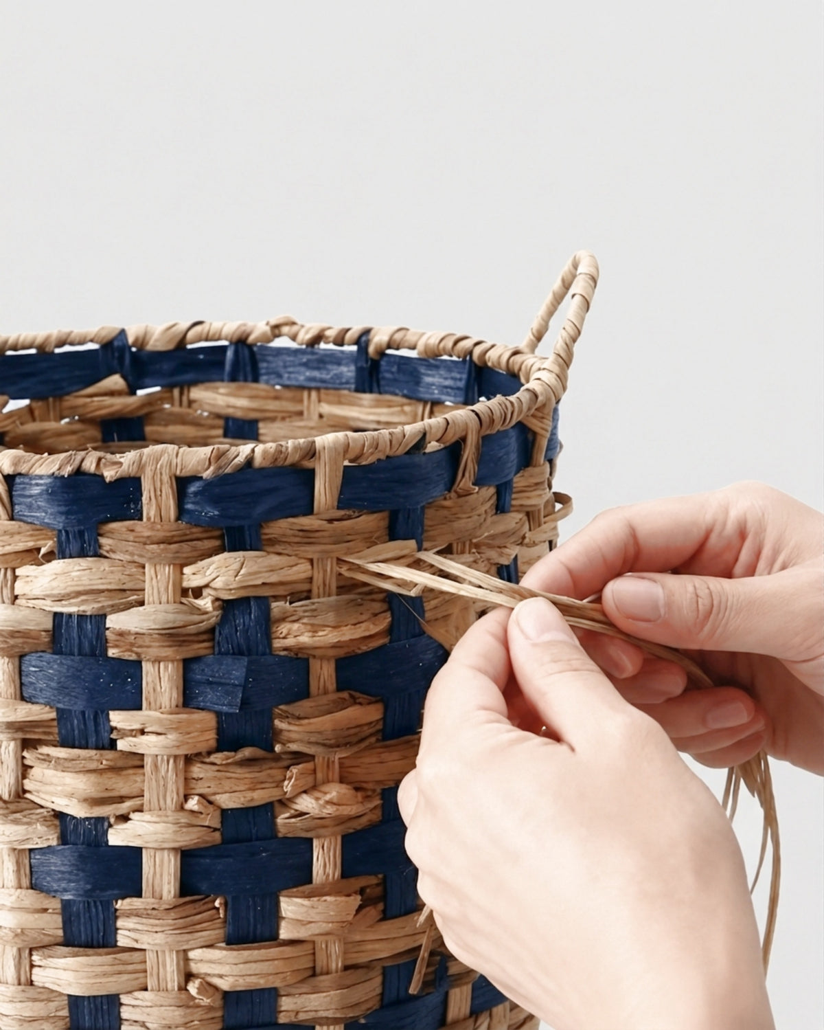 Hands weaving natural grass strands into the deep navy and natural two-tone pattern of Iris basket