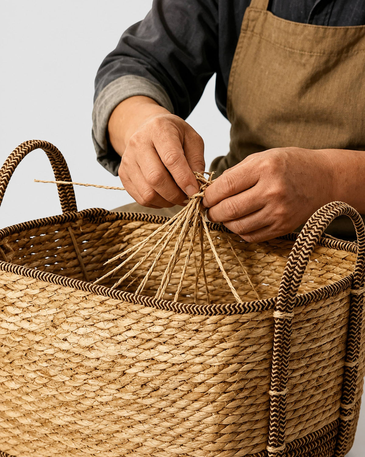 Artisan handweaving natural grass strands into the Vera clothes basket beside its chevron-trimmed handle